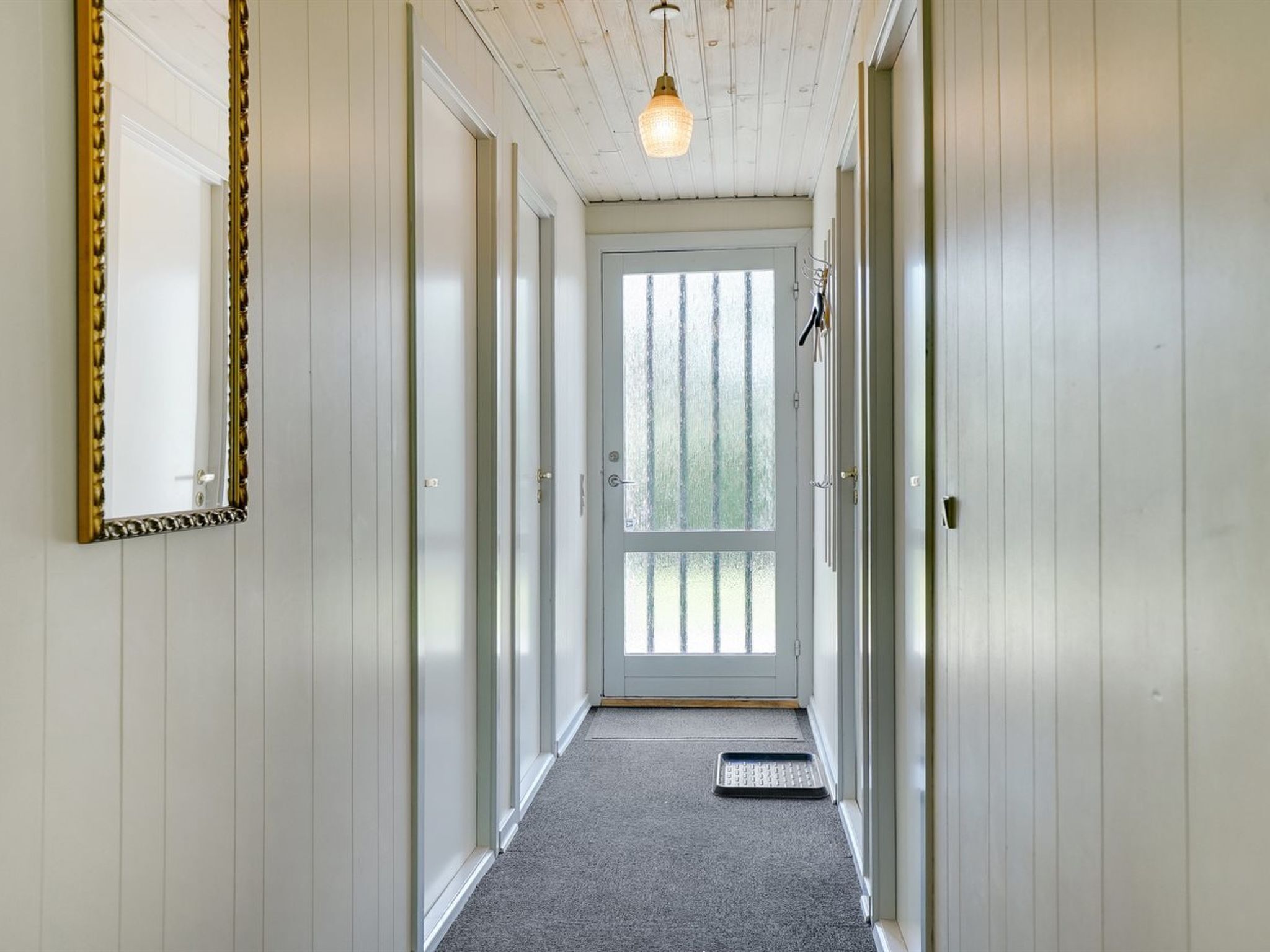 A hallway with white wooden walls, a carpeted floor, a door with frosted glass, and a hanging light fixture.