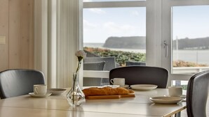 A dining table with a vase of flowers, a croissant, and a view of the sea through large windows.