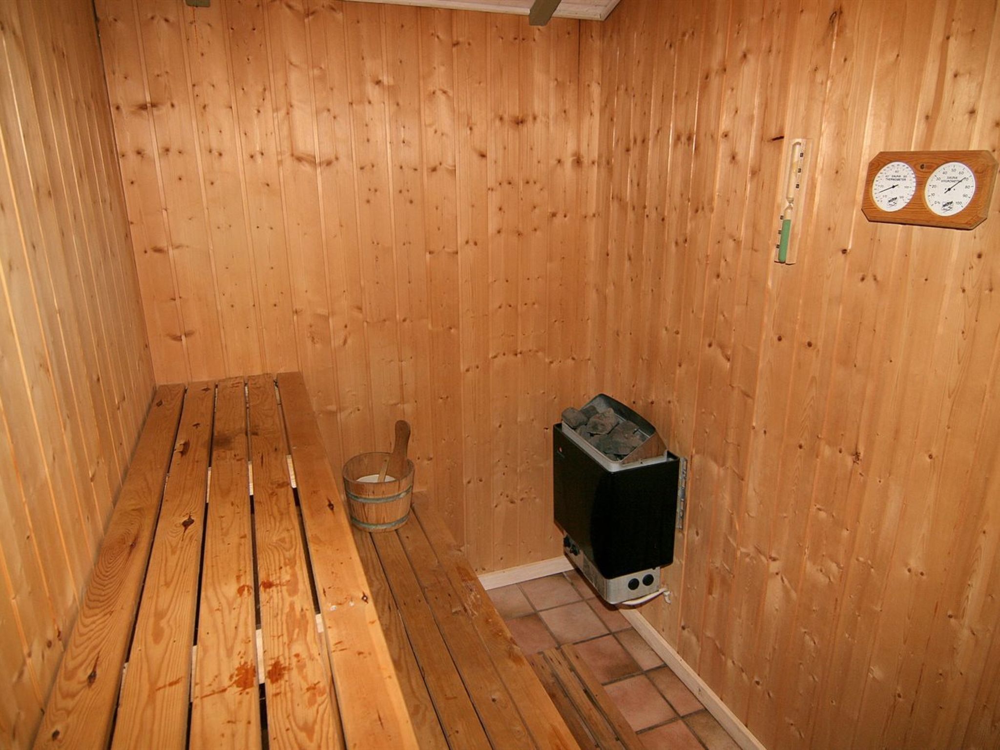A sauna room with wooden walls, a bench, a heater, and a thermometer.