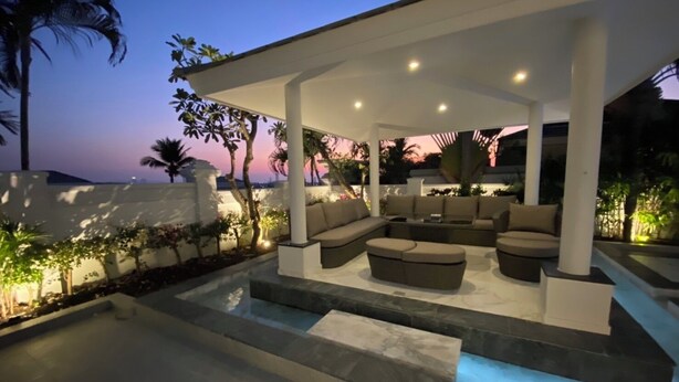 Un área de estar al aire libre techada con piscina, rodeada de palmeras y vista al atardecer.