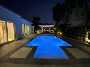Ein Swimmingpool mit blau beleuchtetem Rand, umgeben von einer Terrasse mit überdachter Sitzgelegenheit und einem Gebäude mit Glaswänden.