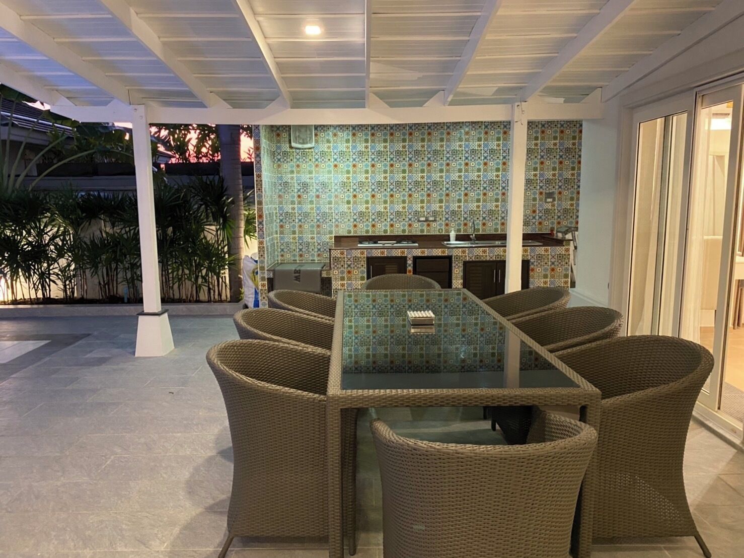 An outdoor dining area with wicker chairs and a glass-top table, surrounded by patterned tiles and a pergola structure.