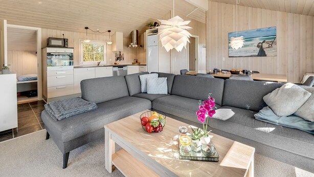 A modern living room with a grey sectional sofa, a wooden coffee table with a fruit bowl and candles, and a kitchen in the background with wooden cabinets and a hanging light fixture.