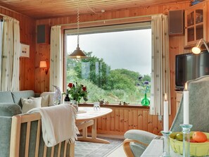 Una habitació acollidora d'estil cabana, amb interior de fusta, un gran finestral que ofereix vistes a la vegetació, taula de menjador amb cadires, televisió i una taula petita amb objectes decoratius.