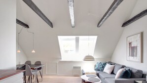 Moderni loft-tyylinen asunto, jossa on näkyvillä olevat kattopalkit, ruokailutila, harmaalla sohvalla varustettu olohuone ja suuri kattovalokuvio.