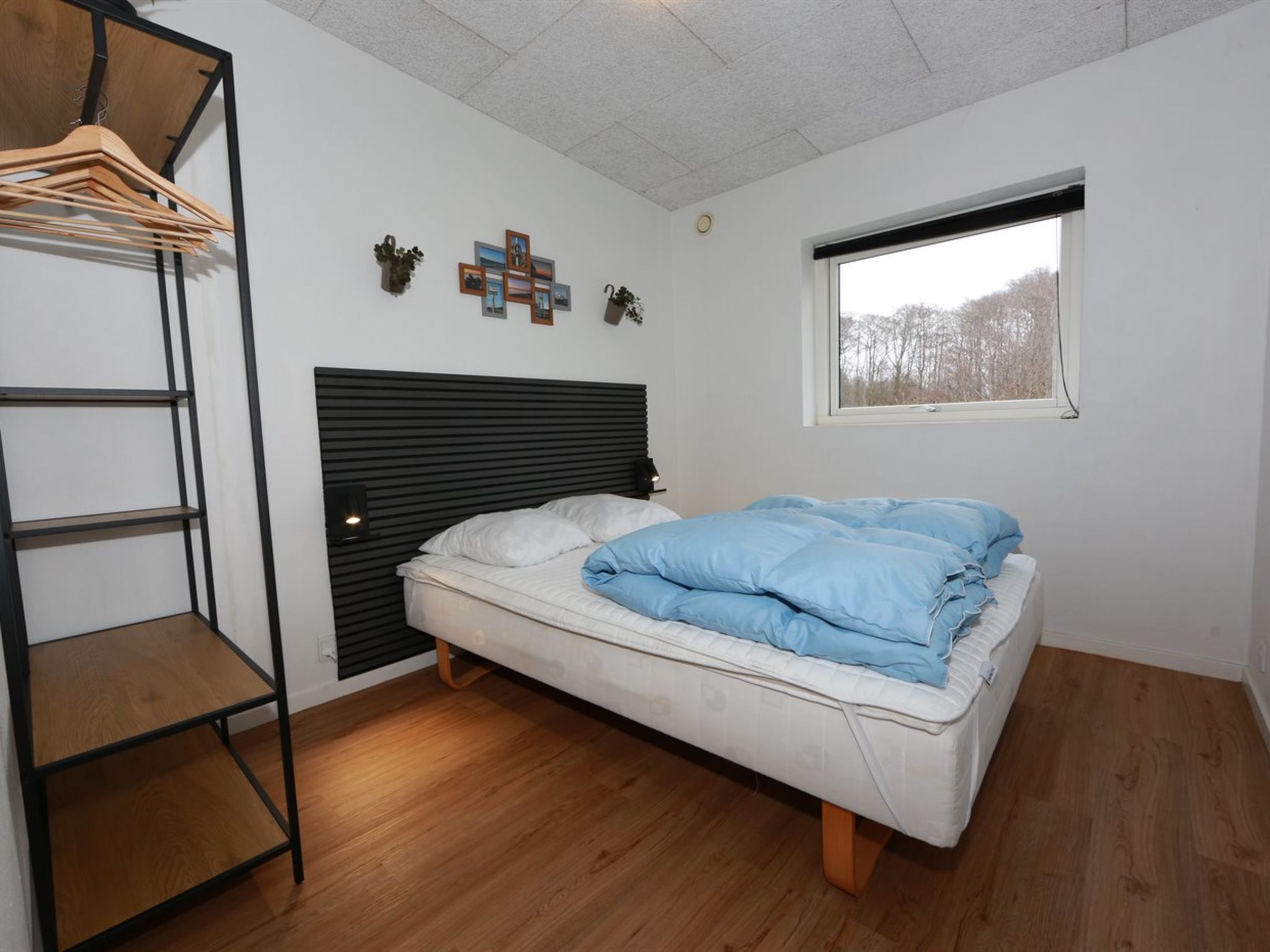 A bedroom with a bed, a wooden shelf, a window, and a wall-mounted picture arrangement.