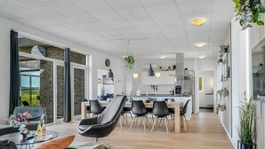 En moderne stue med sofa, lænestole og et spiseområde med bord og stole. I baggrunden er der et køkken med skabe og hårde hvidevarer.
