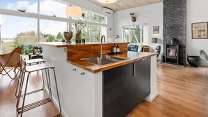 A modern kitchen with a large island, bar stools, a stone fireplace, and large windows offering a view of the outdoors. - "Tenna" - 250m to the fjord by Interhome (Højslev)