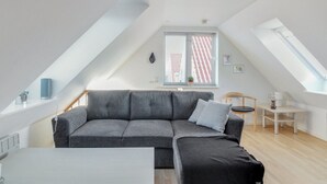 Een modern loft appartement met een grijze hoekbank, een klein houten tafeltje en een groot dakraam.