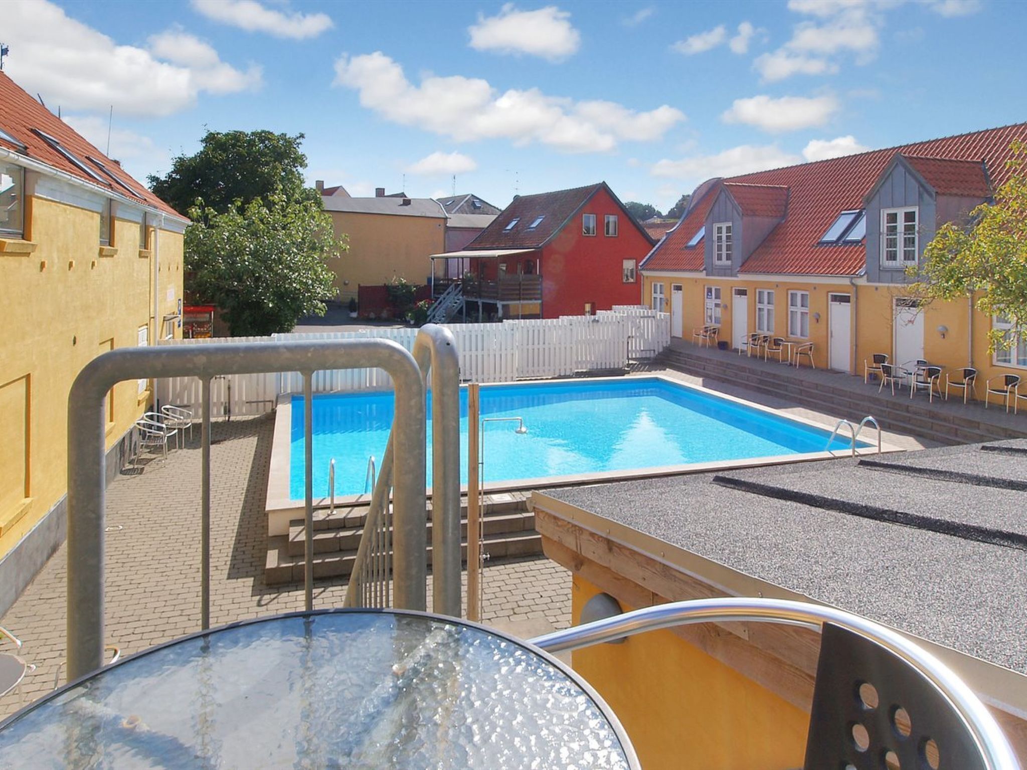 Una piscina con una escalera de metal, rodeada por una valla blanca, y edificios al fondo.