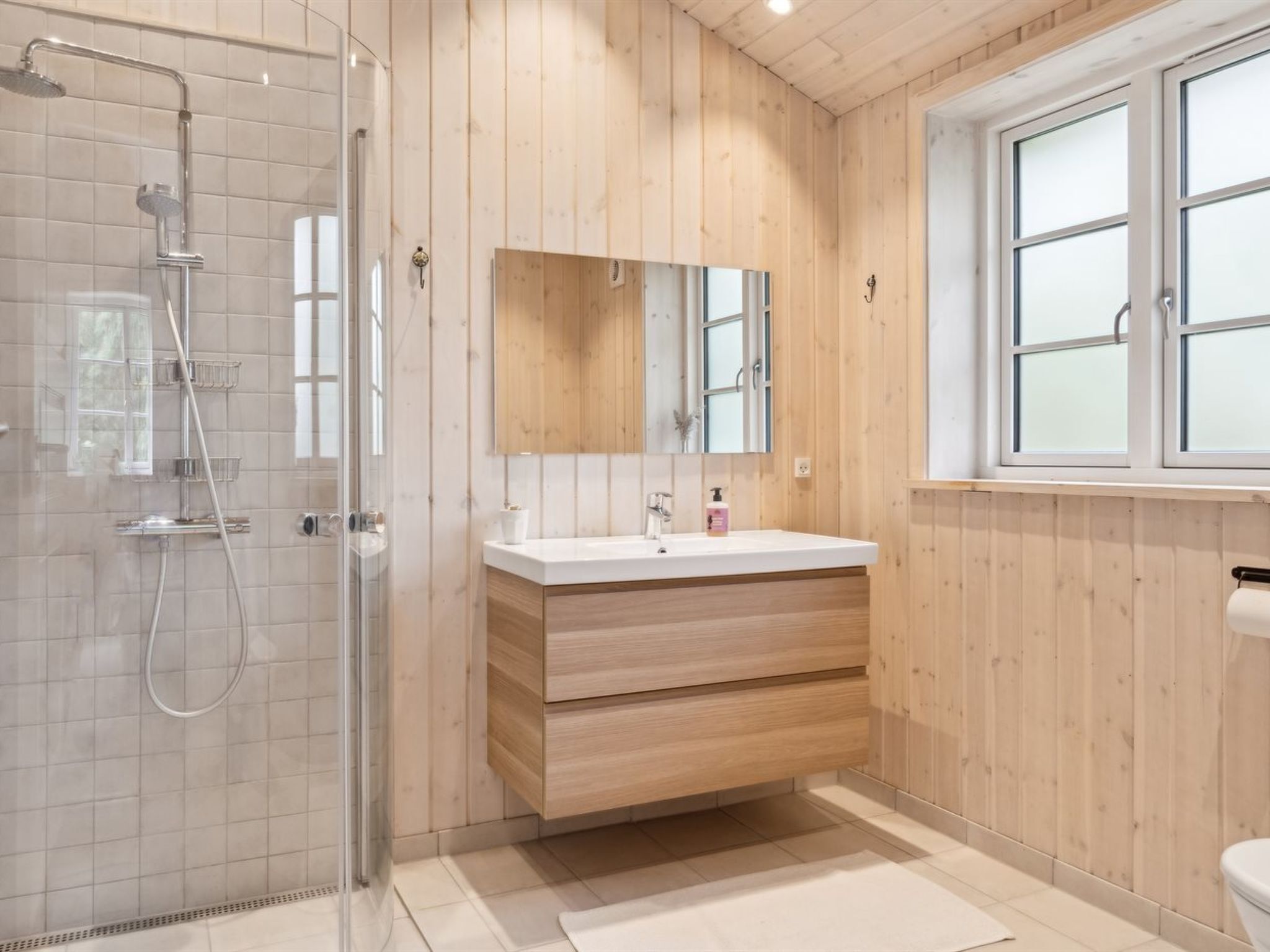A modern bathroom with a glass shower enclosure, wooden panel walls, a white vanity with a mirror, and a window providing natural light.