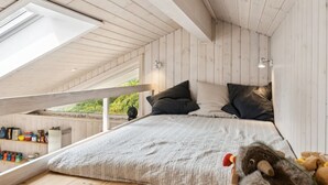 Dormitorio estilo loft con techo inclinado, vigas de madera, cama con colcha gris, almohadas y un peluche.
