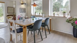 Un comedor con mesa de madera, sillas grises y una lámpara colgante. Hay plantas en macetas en el alféizar de la ventana y una silla de mimbre con una manta de piel.