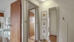 Un pasillo con suelo de madera, paredes blancas y puertas que dan a otras habitaciones. En la esquina, hay un banco blanco y una planta en maceta.