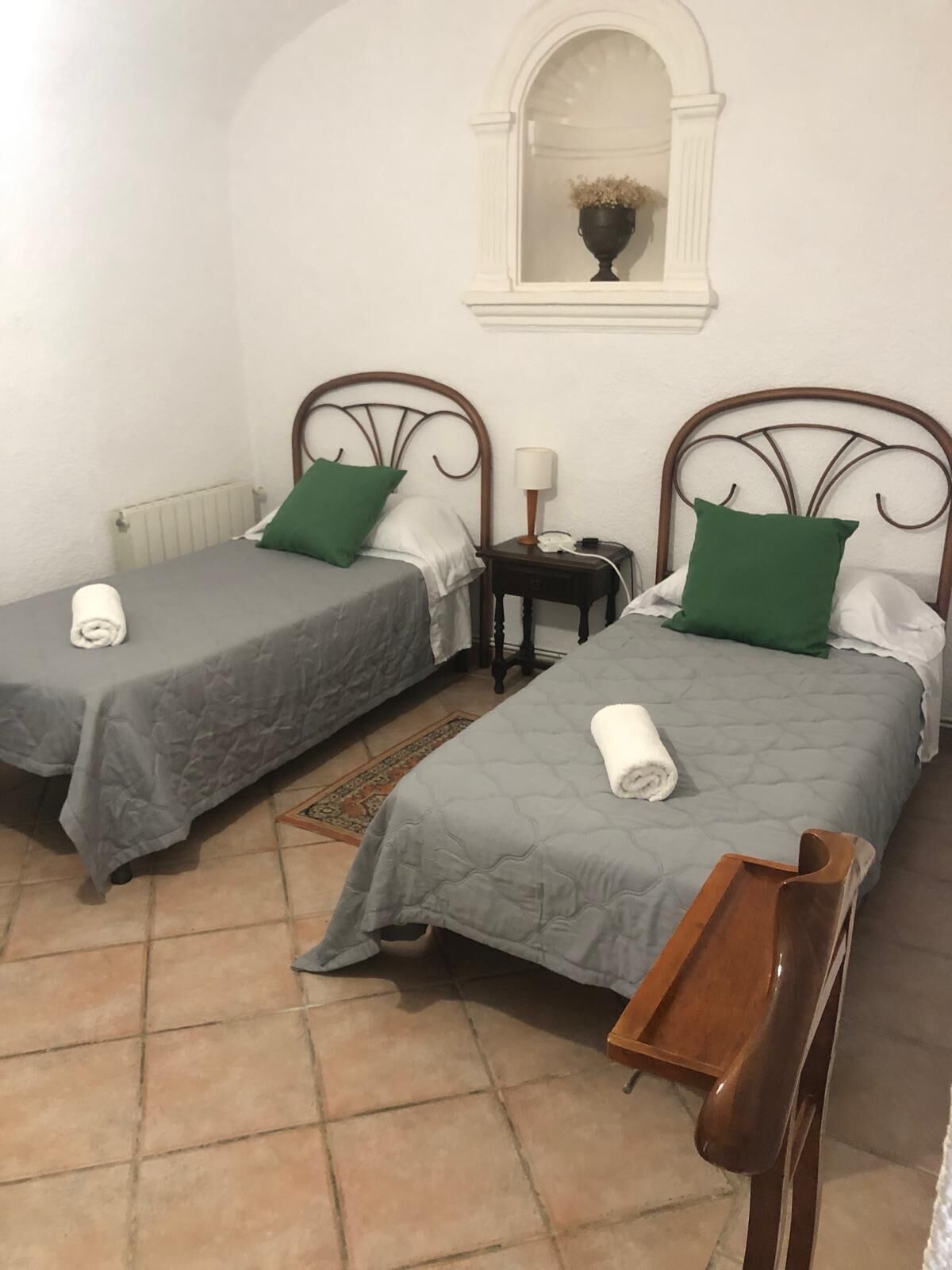 Two single beds with grey bedding and green pillows, a small bedside table with a lamp, and a wooden chair.