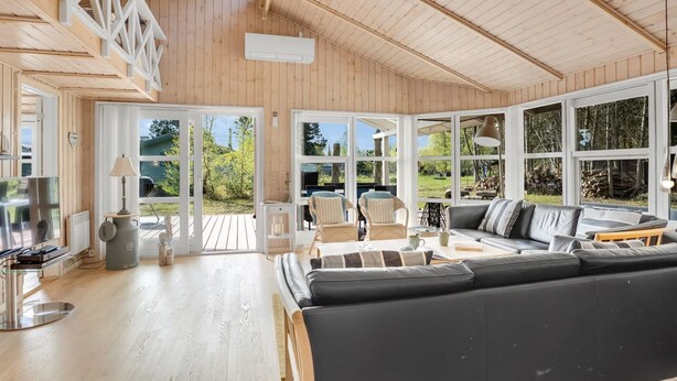 A spacious living area with a large black sofa, wicker chairs, and a wooden deck visible through large windows.