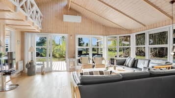 Un vaste espace de vie comprenant un grand canapé noir, des fauteuils en osier et une terrasse en bois visible à travers de grandes fenêtres.