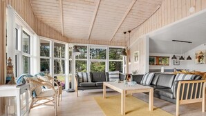 A spacious living room with a wooden ceiling, large windows, a sofa, a coffee table, and a dining area.