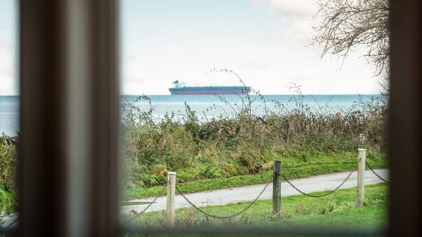 A view of a large ship on the water, a pathway with a chain-link fence, and some vegetation.