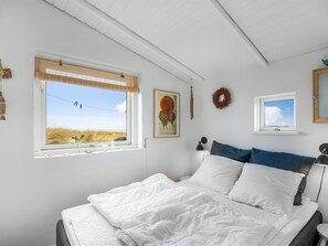 A bedroom with a large bed, a window with a view of the beach, a framed picture, and a small window with a view of the sky.
