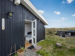 Un bâtiment moderne de couleur noire, doté d’une terrasse en bois, d’une entrée avec une porte en verre et d’un petit espace extérieur aménagé pour s’asseoir.