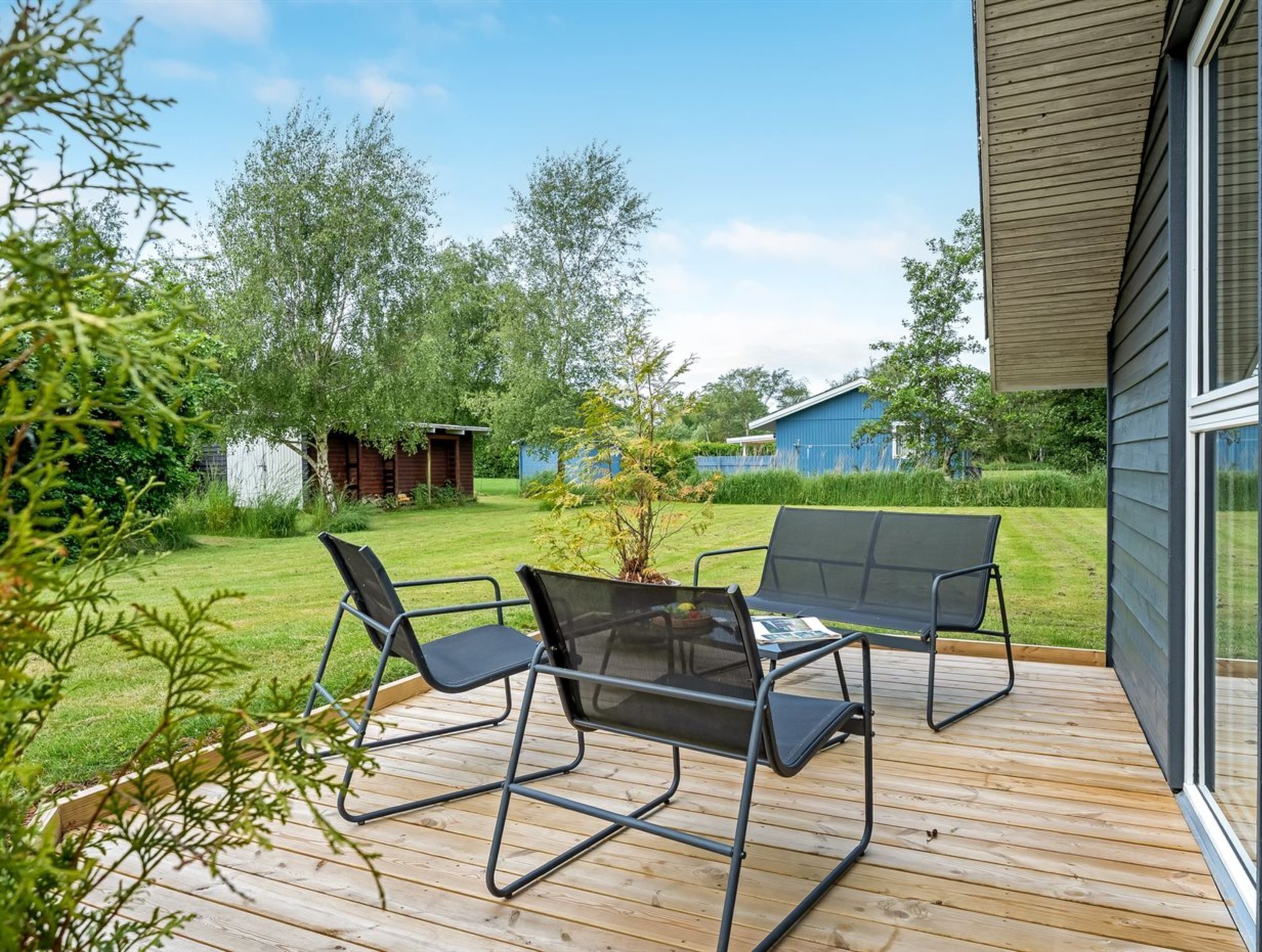 Una terraza de madera con dos sillas negras y una mesa, rodeada de un jardín con césped y árboles.