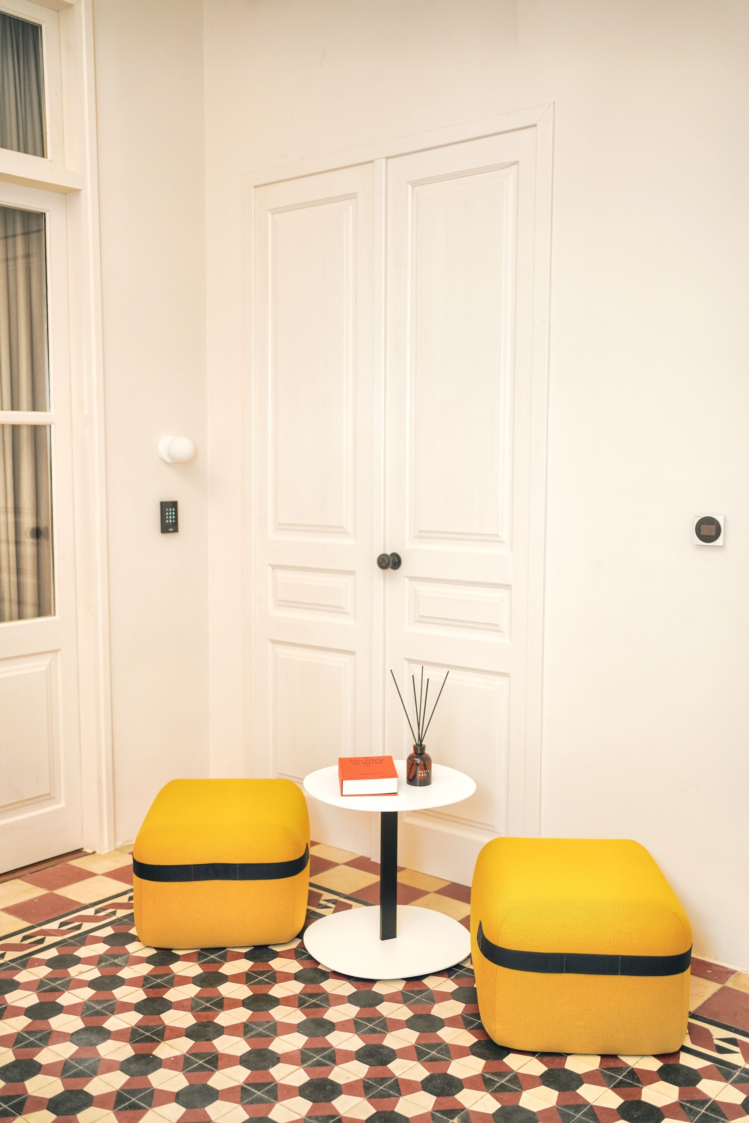 A room with a white door, a small round table with a vase, and two yellow ottomans on a patterned floor.