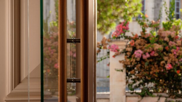 Eine Glastür mit Metallgriff, die zu einem Garten mit rosa Blumen und einem Gebäude im Hintergrund führt.