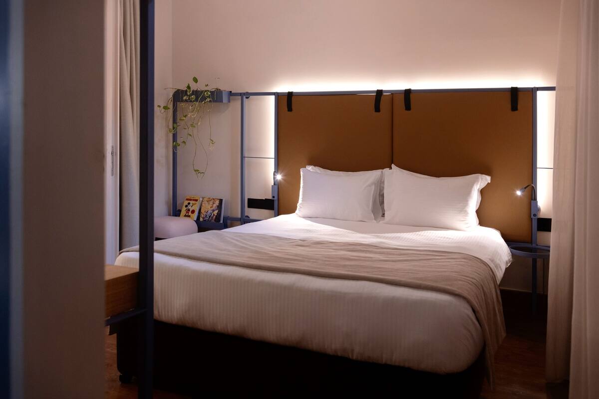 A neatly made bed with white linens, a bedside table with a lamp, and a wall-mounted shelf with a plant.