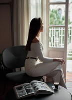 Uma pessoa sentada em uma cadeira perto de uma janela, com um livro e uma xícara sobre a mesa.
