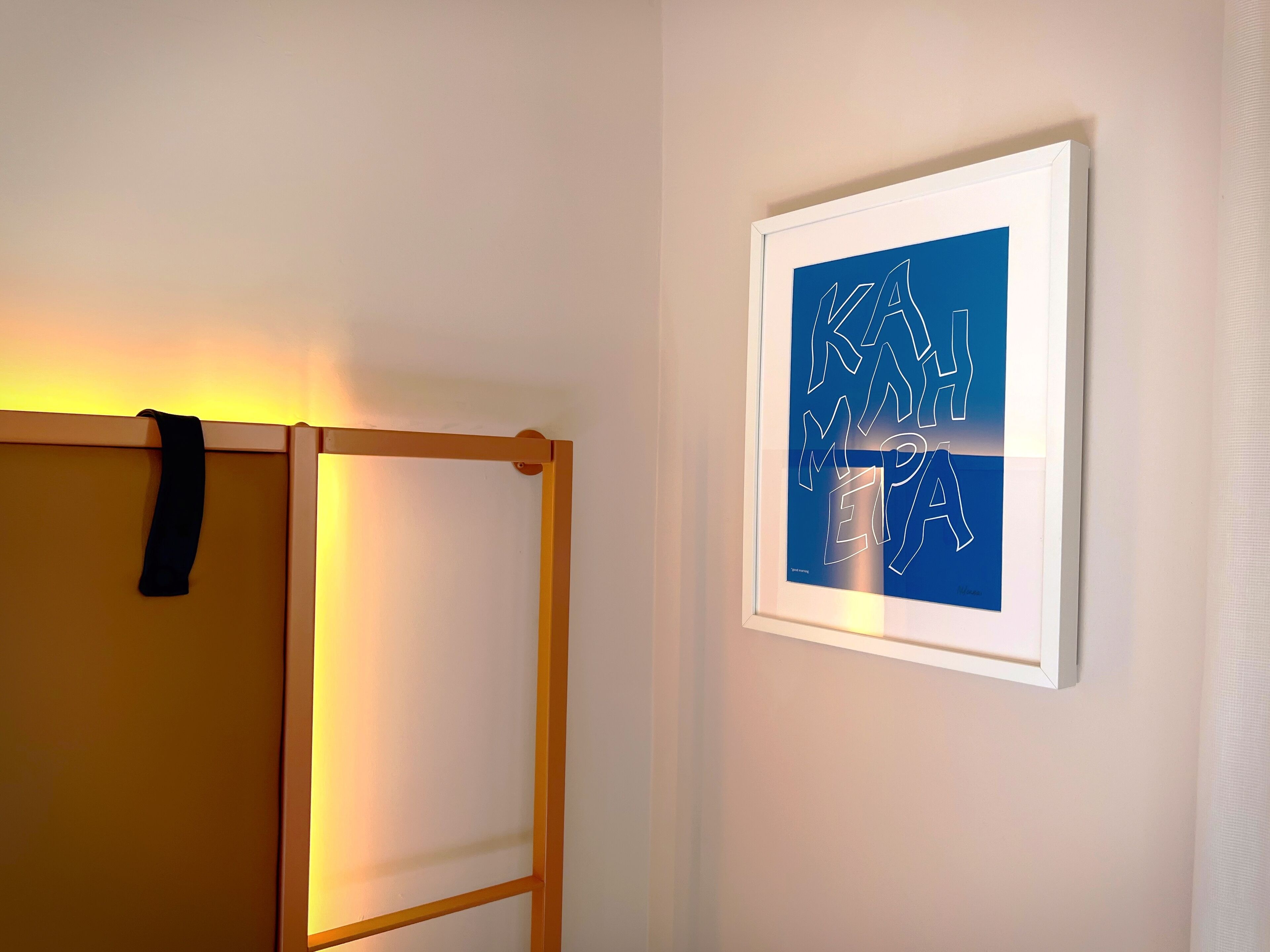 Ett inramat konstverk på en vägg, en träkonstruktion med ett svart band och en ljuskälla till vänster.