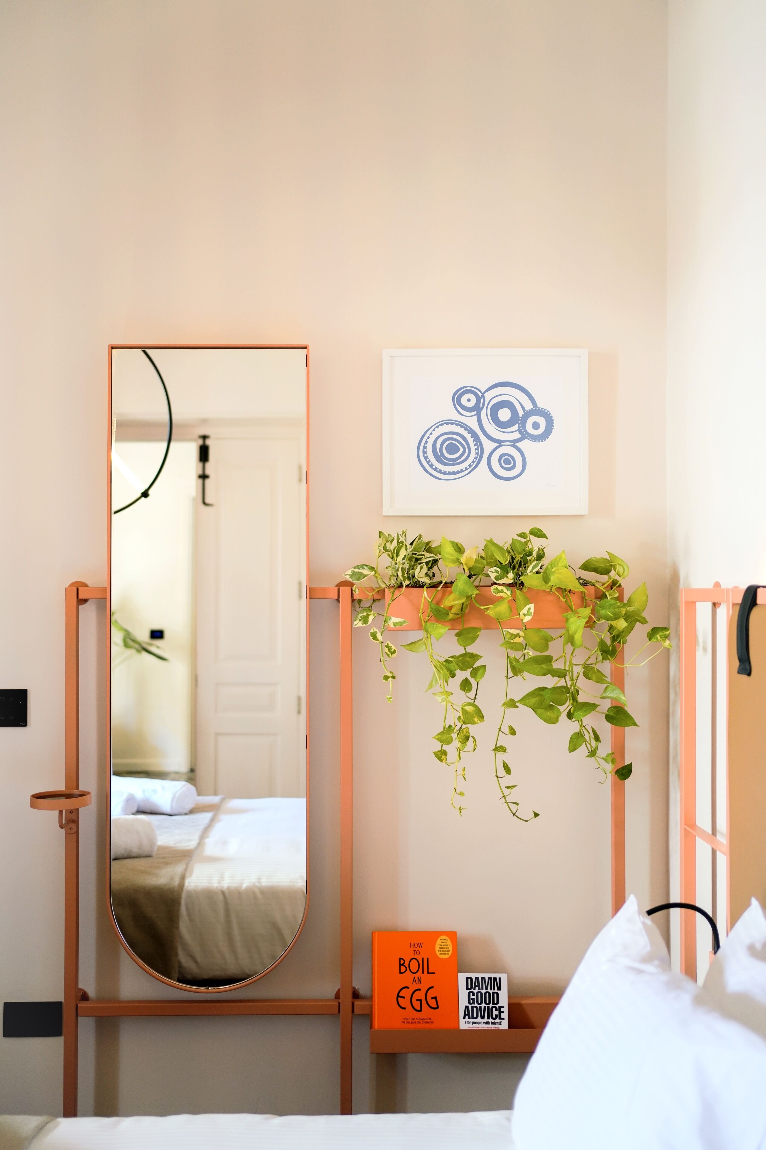 A bedroom with a large mirror, a wall-mounted plant shelf, a framed abstract artwork, and a bedside table with a book titled "How to Boil an Egg."