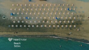 Eine Vogelperspektive zeigt einen Strand mit zahlreichen runden Strukturen, möglicherweise Zelten oder Hütten, die in Reihen angeordnet sind. Neben dem Strand erstreckt sich ein klares, blaues Meer, und in der Ferne sind einige Personen zu sehen. Der Text 'hearthotel Beach 500m' gibt den Standort an.