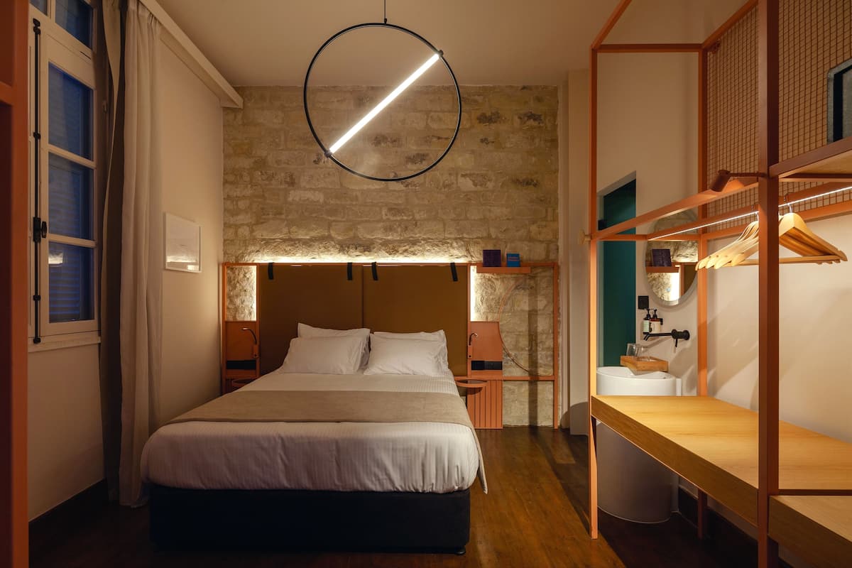 A bedroom with a large bed, a hanging circular light fixture, a stone wall, and a wooden bench.
