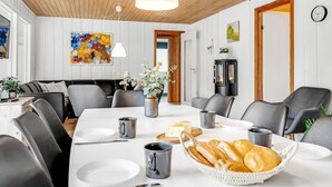 A dining area with a wooden ceiling, white walls, and a black leather couch. There is a table set with plates, cutlery, and a basket of bread. - "Sverker" - 300m from the sea by Interhome (Rødby)