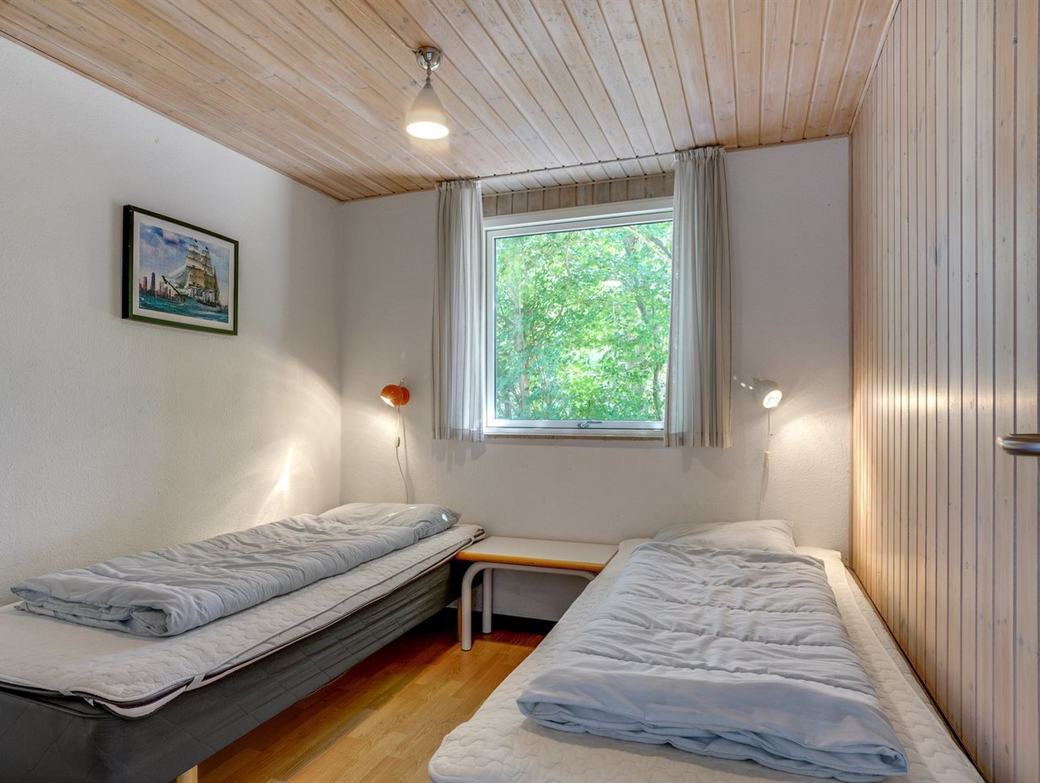 Et lille værelse med to enkeltsenge, et vindue med gardiner, en lampe på væggen og et billede i ramme.