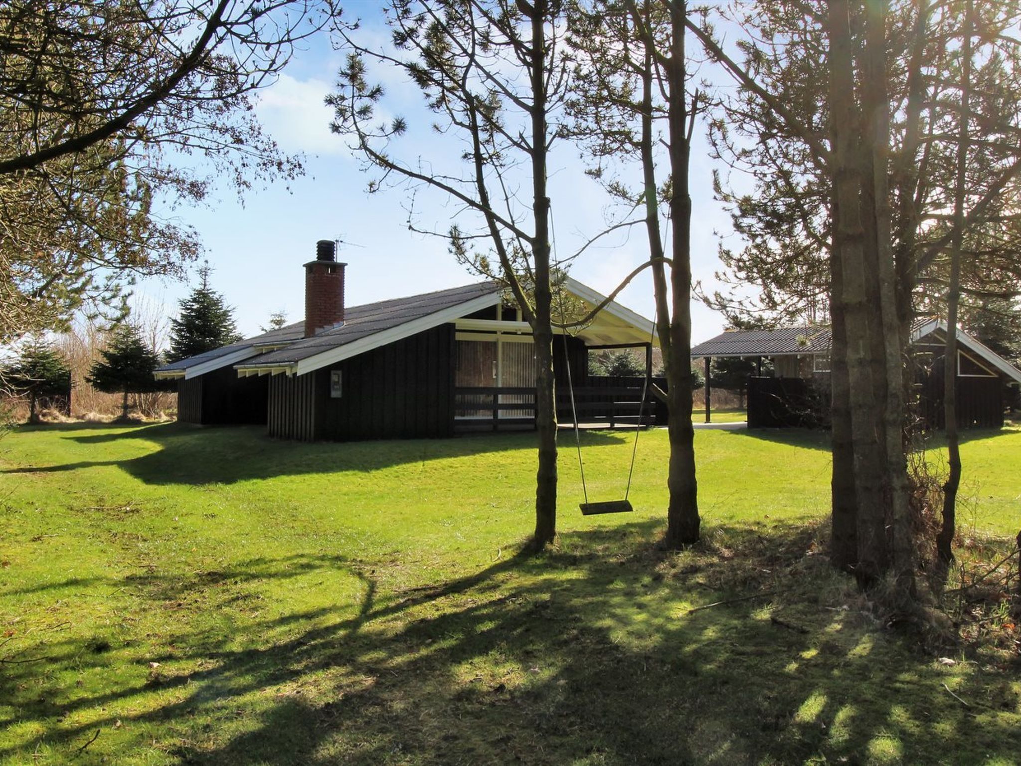 A rustic cabin with a sloped roof, a chimney, and a swing set in a grassy yard surrounded by trees.