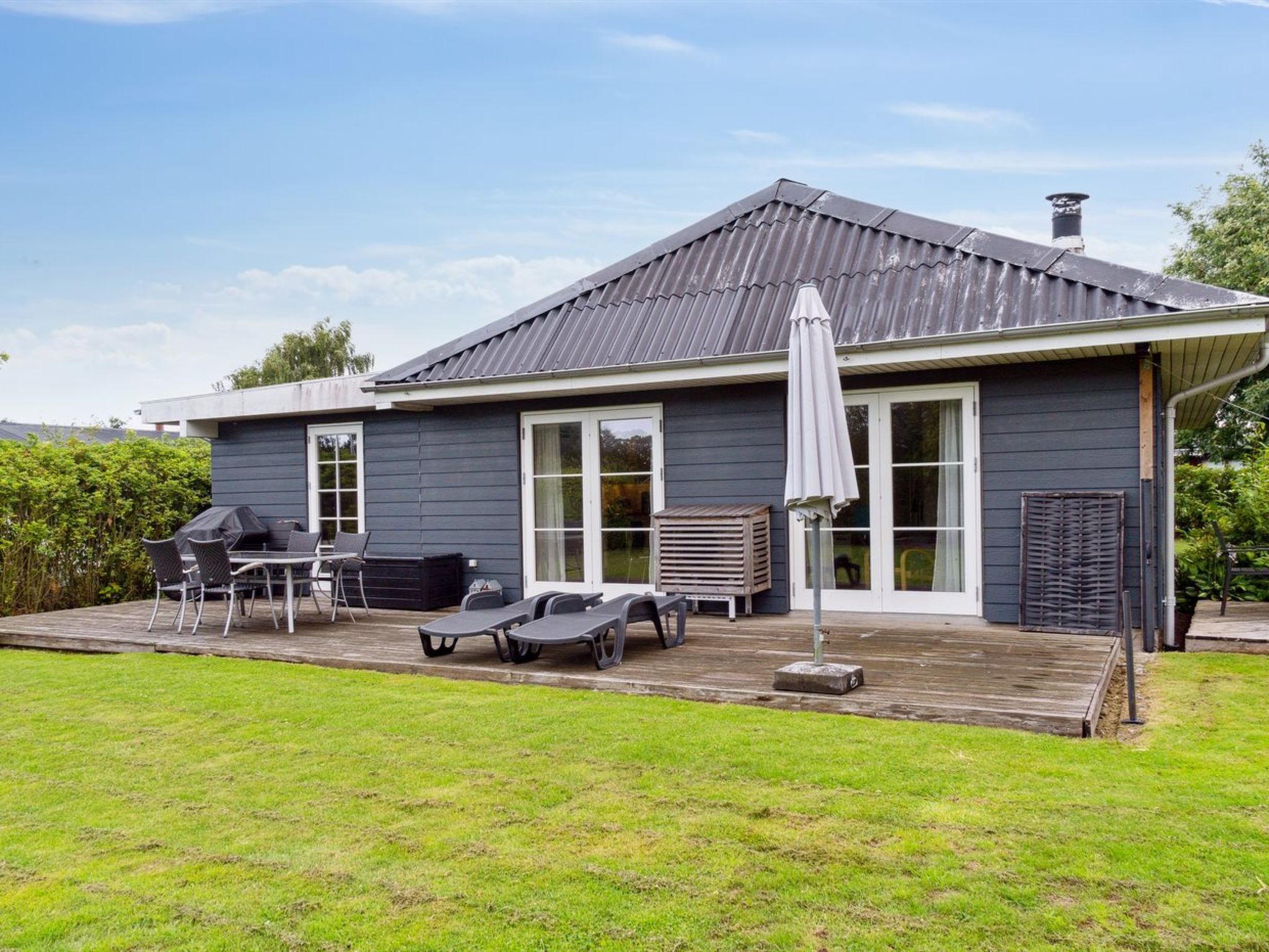 Een modern huis met een donkergrijze gevel, een schuin dak en een overdekte patio met buitenmeubilair en een barbecue.