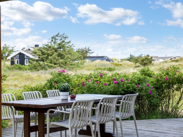 A wooden deck with a table and chairs, surrounded by greenery and pink flowers.