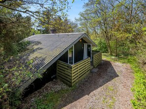 Eine kleine Holzhütte mit geneigtem Dach, einer Tür und einem Fenster, umgeben von Bäumen und einem Kiesweg.