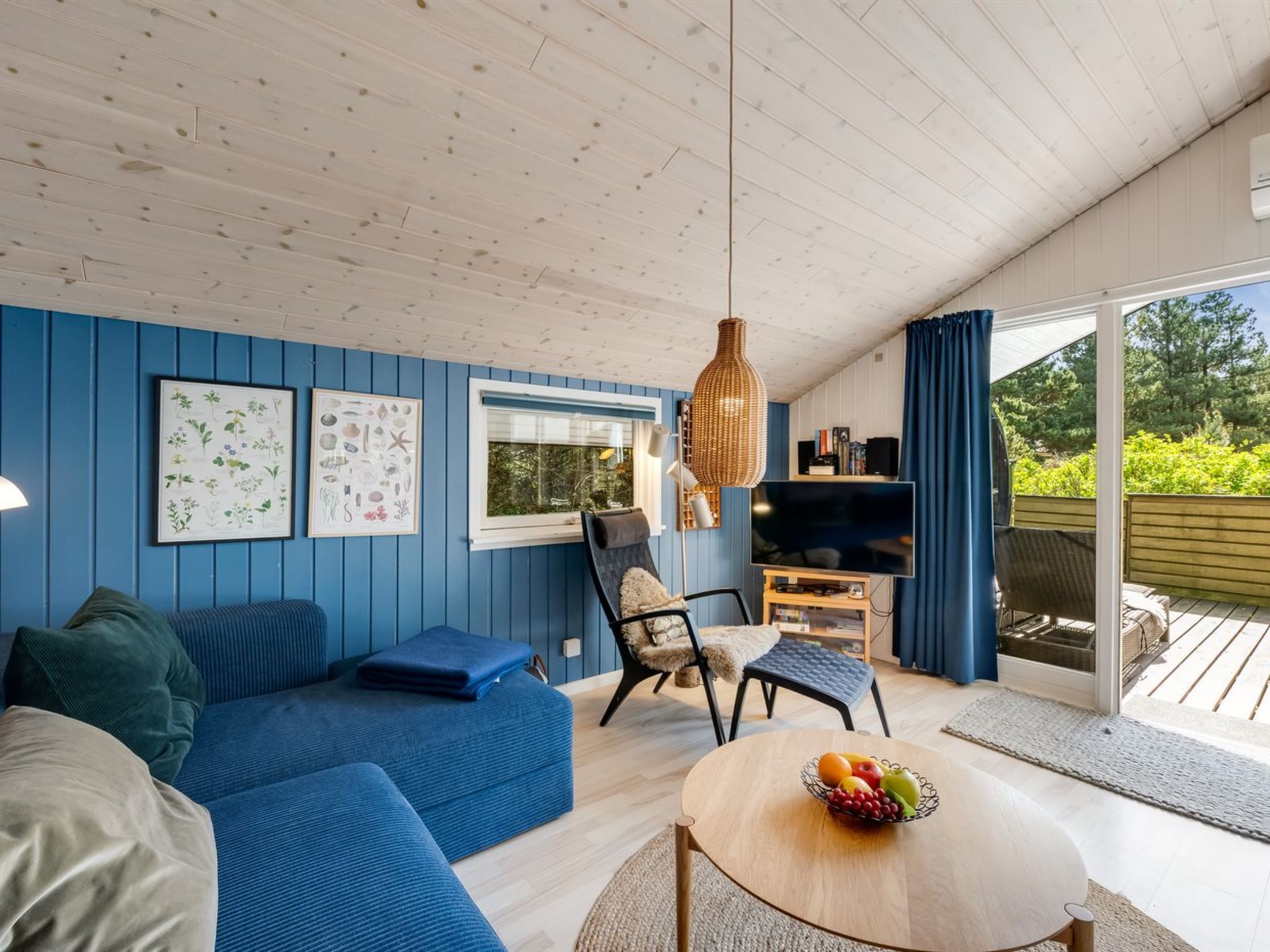 Een kamer met een blauwe wand, een houten plafond, een bank, een stoel, een tafel met fruit en een televisie.