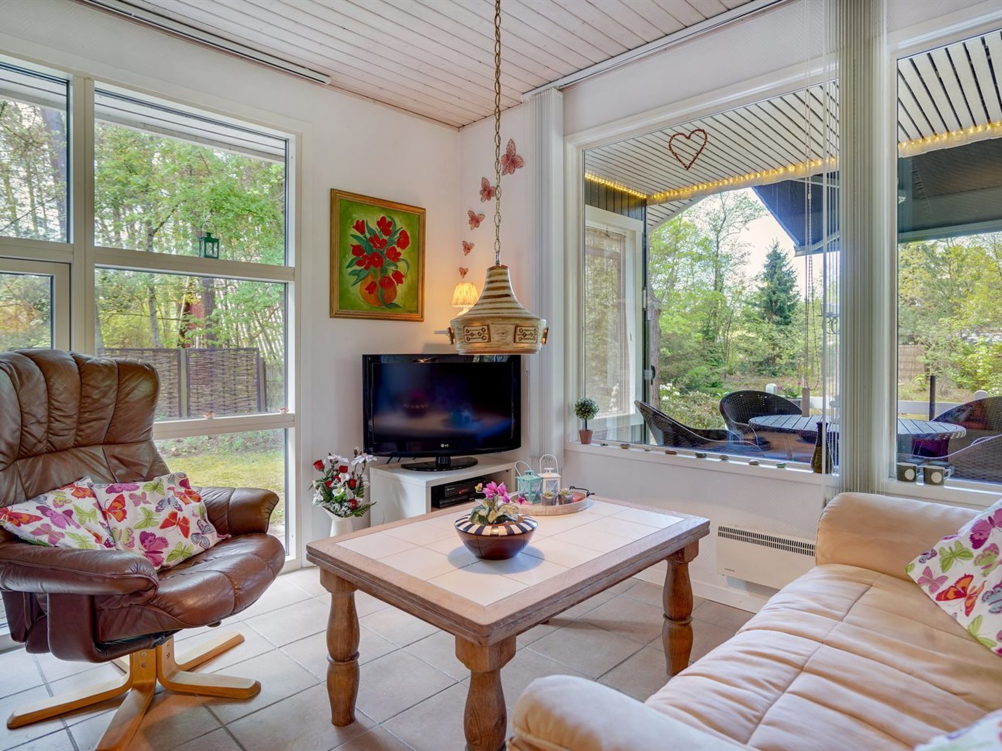 Un salon chaleureux avec un canapé en cuir, une table basse en bois et un téléviseur. De grandes fenêtres offrent une vue sur l’extérieur.