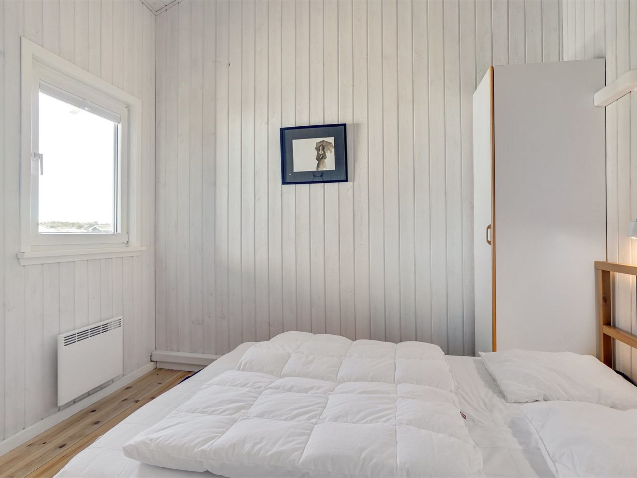 Une chambre avec un grand lit, une fenêtre, un radiateur et un tableau encadré accroché au mur.