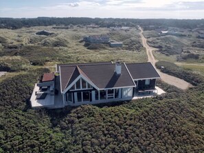 An aerial view of a modern house with a sloped roof, surrounded by dense vegetation and a dirt road.