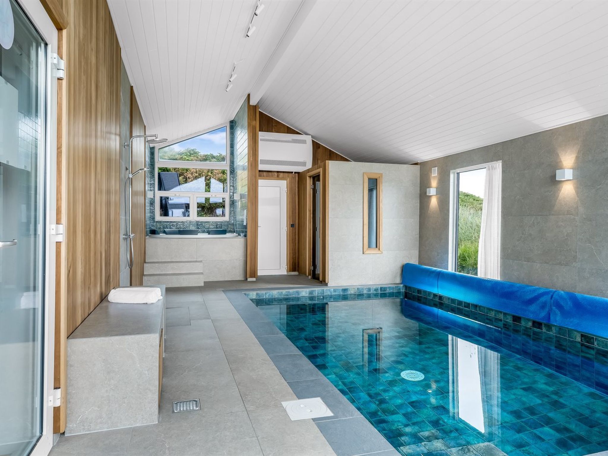 A modern indoor swimming pool with clear blue tiles, a seating area, and a view of the outdoors.