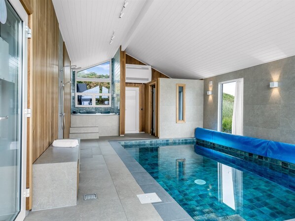 A modern indoor swimming pool with clear blue tiles, a seating area, and a view of the outdoors.
