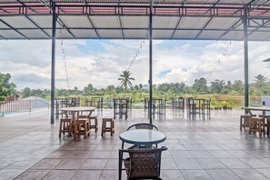 Een buitenterras met tafels en stoelen, een overdekt gedeelte en uitzicht op palmbomen en groen.