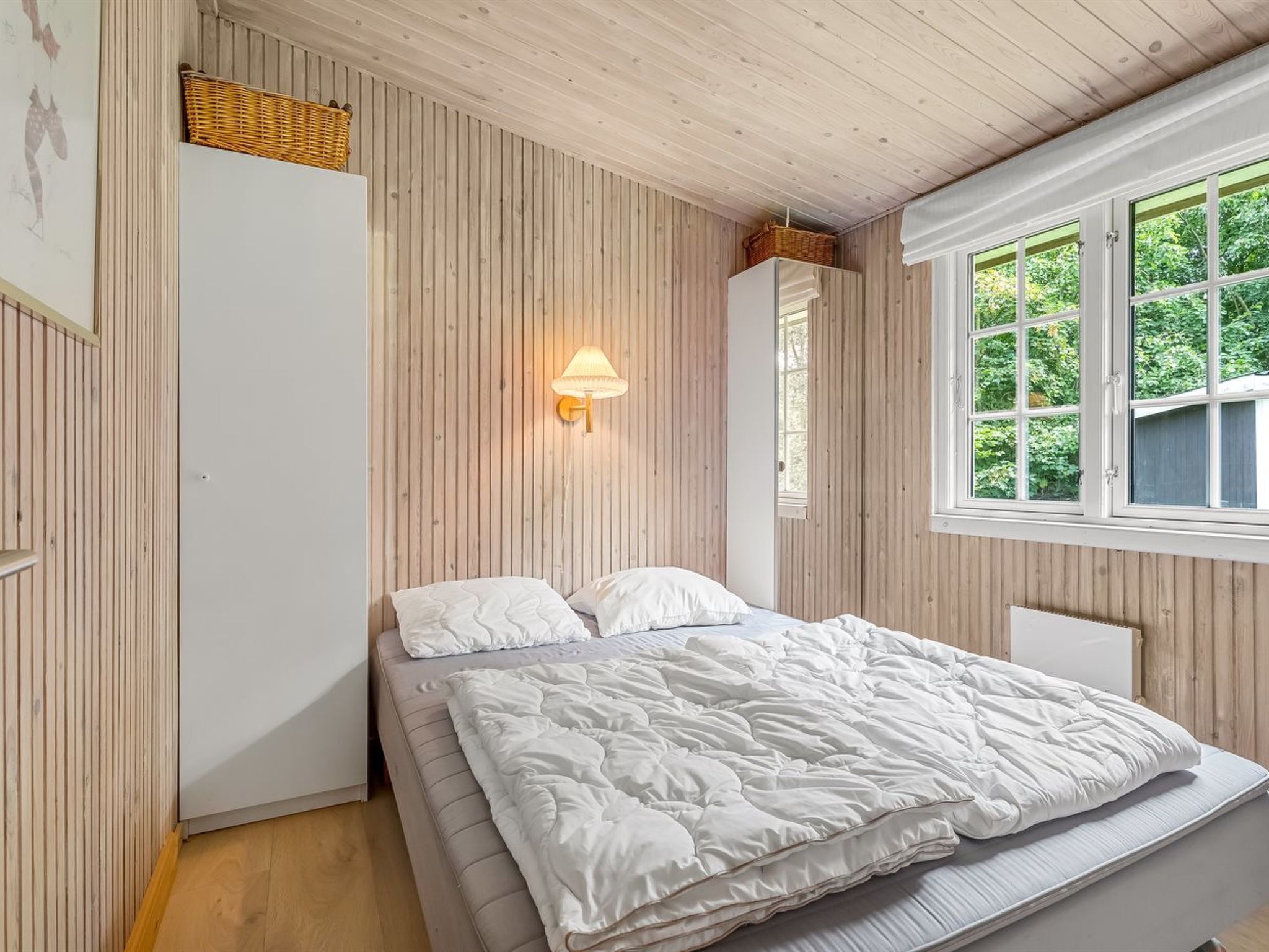 Ein Schlafzimmer mit einem Bett, einer Wandleuchte, einem Fenster mit Blick ins Grüne und einer Holzbalkendecke.