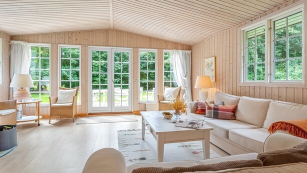 Una habitación acogedora con paredes de madera y ventanas amplias, un sofá blanco, una mesa de centro y sillas de mimbre.