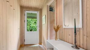Un pasillo estrecho con paredes de madera, una puerta blanca con un ventanal verde, una consola blanca con un jarrón de plantas secas y un espejo en la pared.